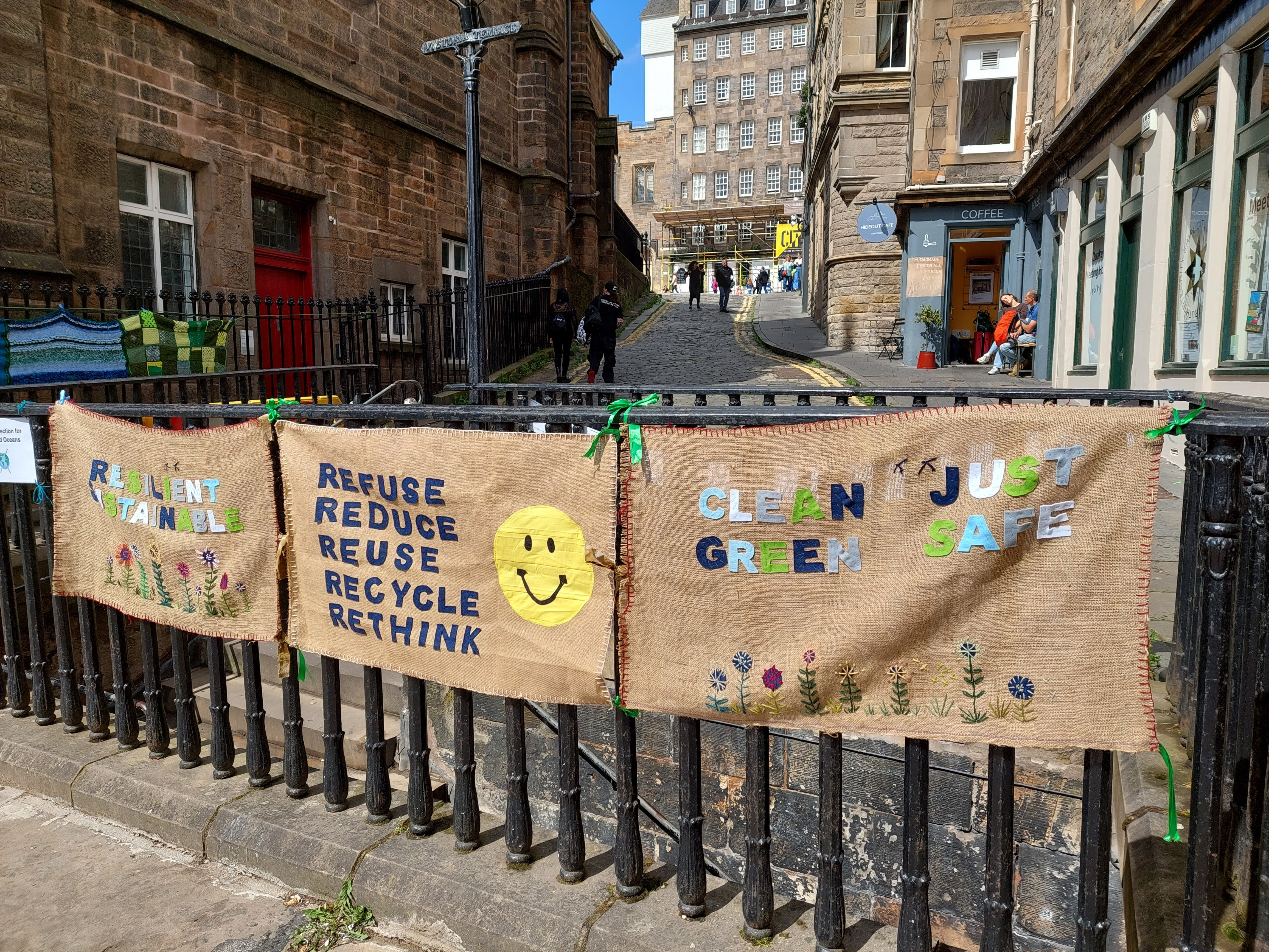Three banners tied to a railing on an Edinburgh Street. They are hessian with words appliqued on. Left it says 