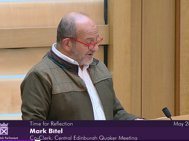 Mark Bitel, Co-Clerk of Central Edinburgh Quaker Meeting, addresses Scottish Parliament. Mark is wearing a white shirt, green jacket and red glasses.