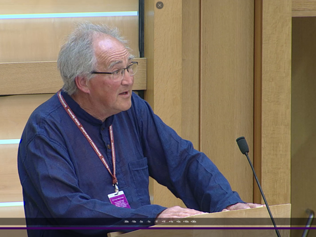 Richard Raggett, a Quaker from North Scotland, speaks to Parliament. Richard has grey hair, glasses, and is wearing a dark blue shirt standing in front of the podium in the Scottish Parliament